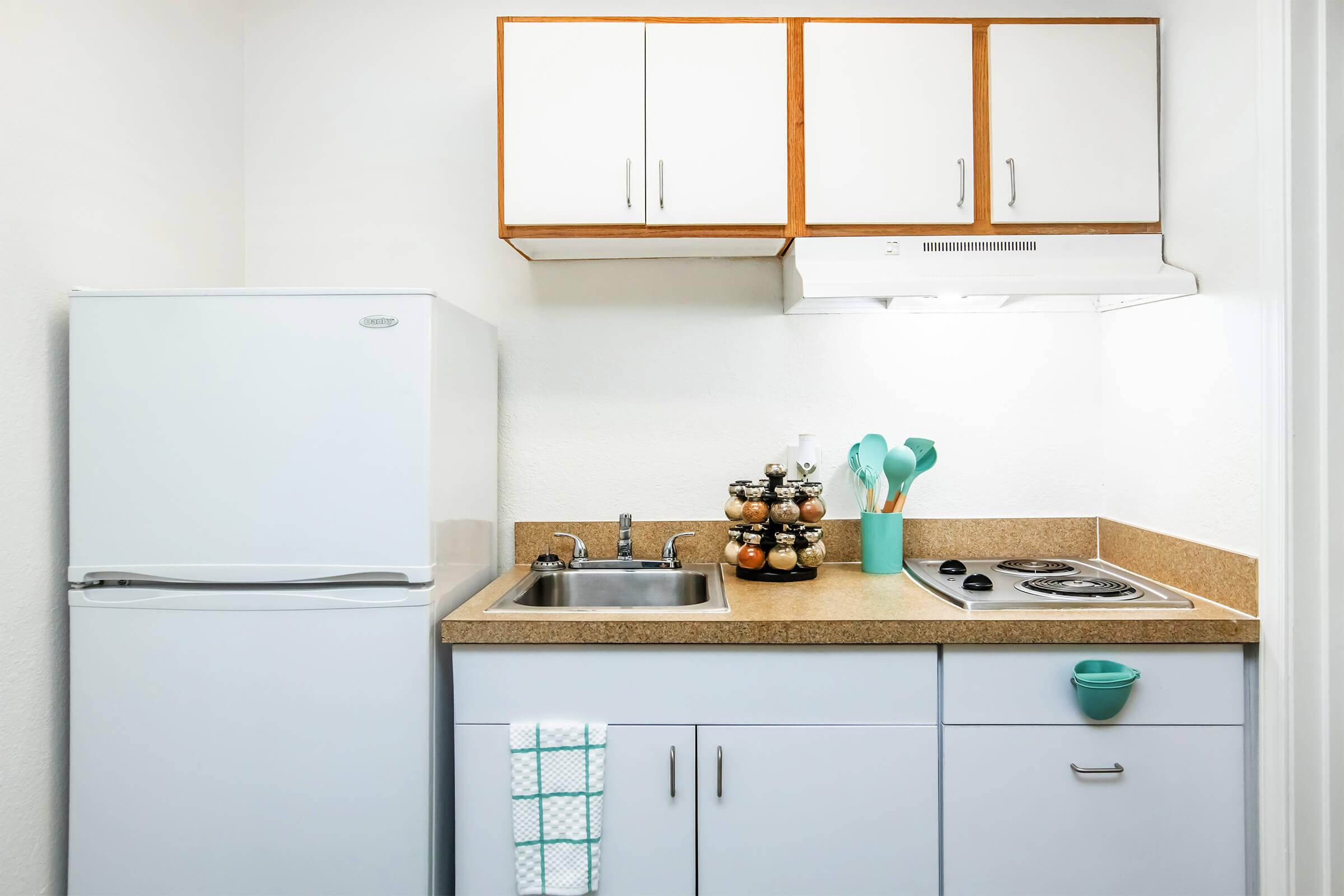 A small kitchen setup featuring a white refrigerator, a sink, and a stovetop. Above, there are white cabinetry and a range hood. A countertop holds a container with cooking utensils and a display of spice jars. A dish towel hangs from the cabinet, and there is a small decorative bowl on the counter.