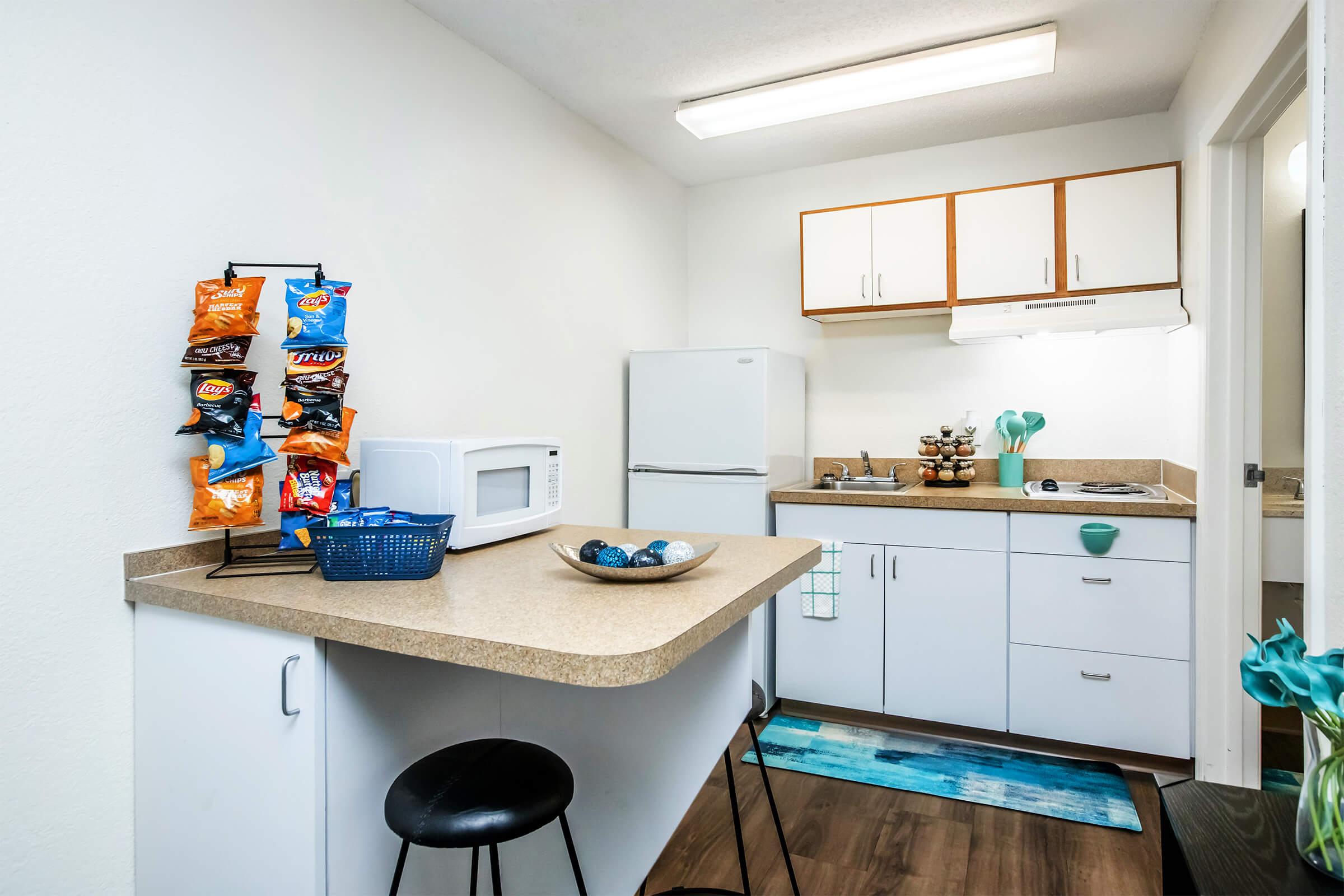 Modern kitchenette featuring a countertop with bar stools, a microwave, and a refrigerator. Snack items are displayed on a rack, and there’s a small bowl of decorative items on the counter. Cabinets are above the sink, and a colorful rug adds ambiance to the space.