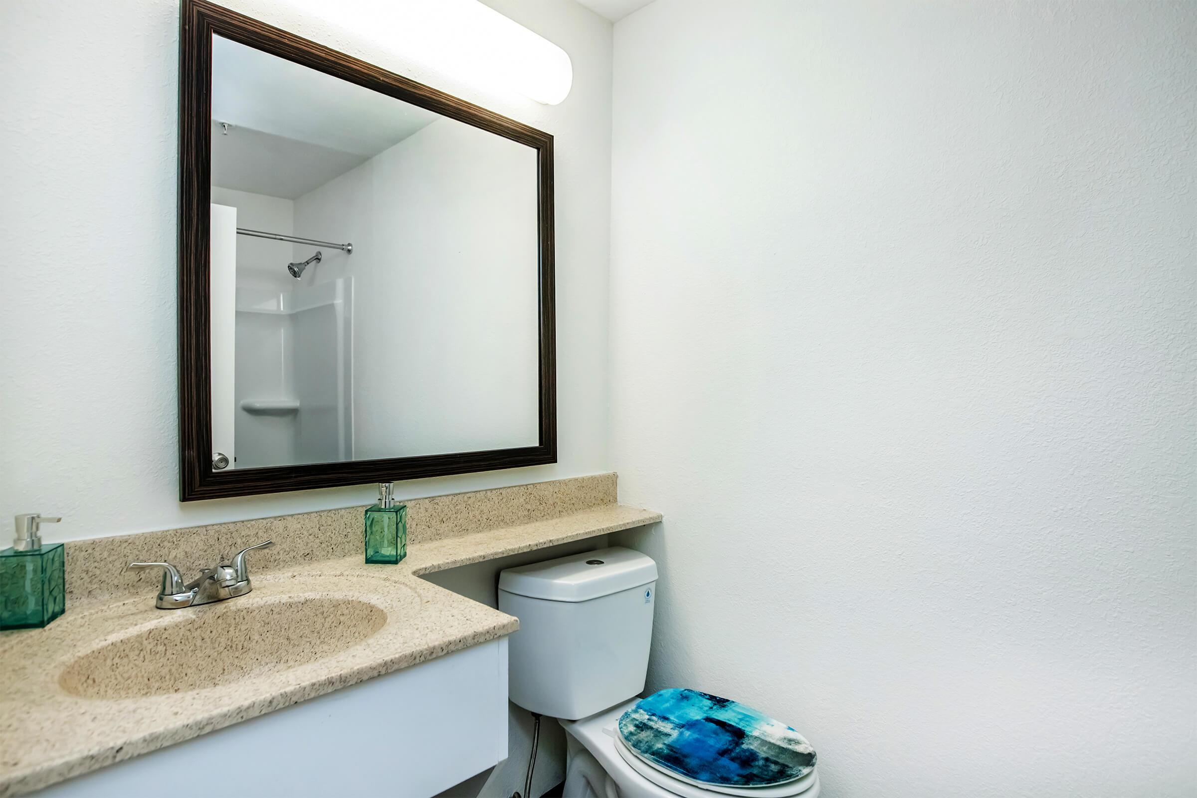 A clean, modern bathroom featuring a mirror above a beige countertop, a toilet with a blue patterned seat cover, and two green glass soap dispensers. There is a shower stall visible in the background, and the walls are painted white, giving the space a bright and airy feel.