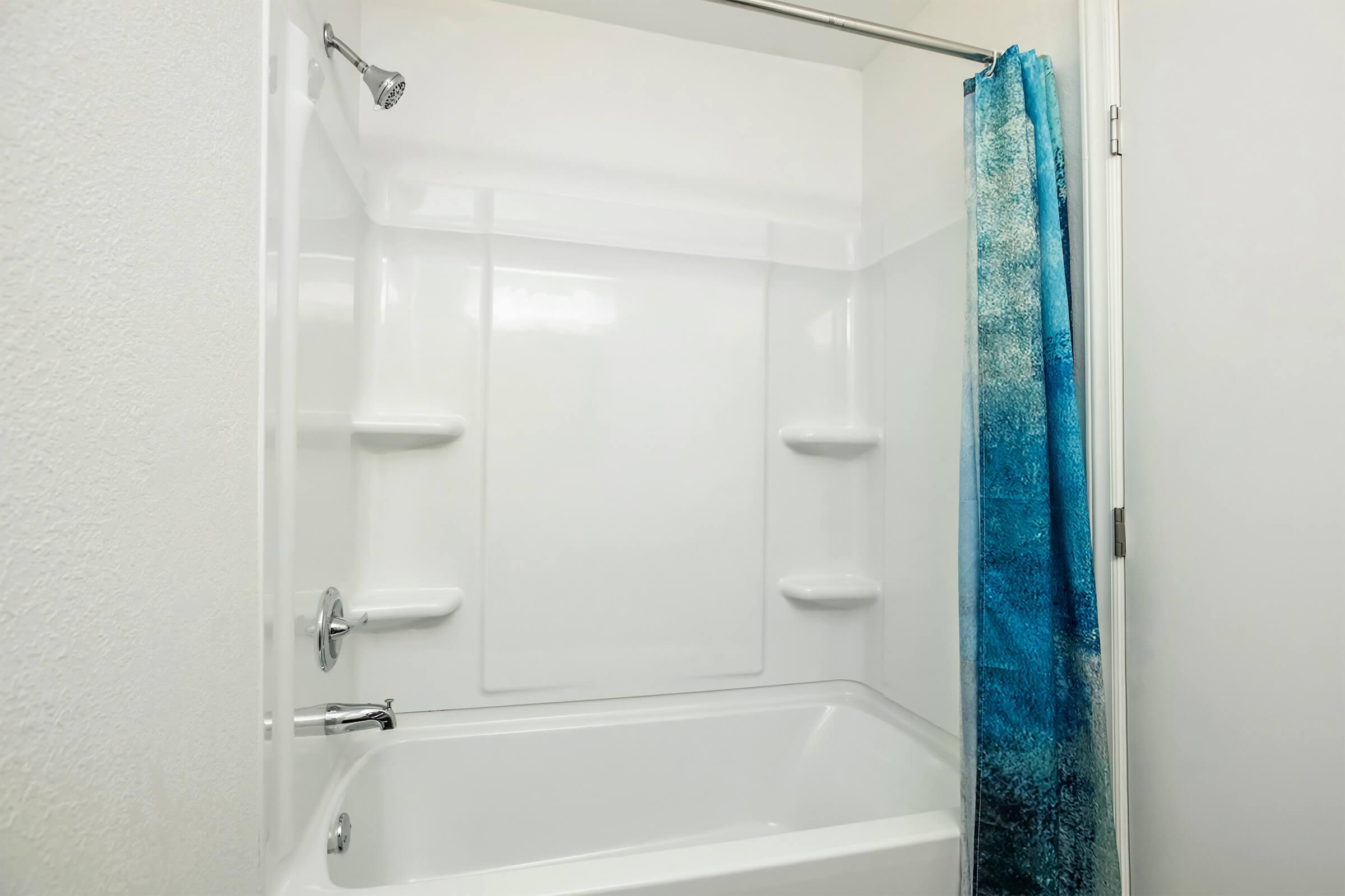 A clean, modern bathroom with a white bathtub and shower. The shower area features a blue and teal gradient shower curtain, and there are built-in shelves for toiletries. The walls are light-colored, enhancing the bright and spacious feel of the room.