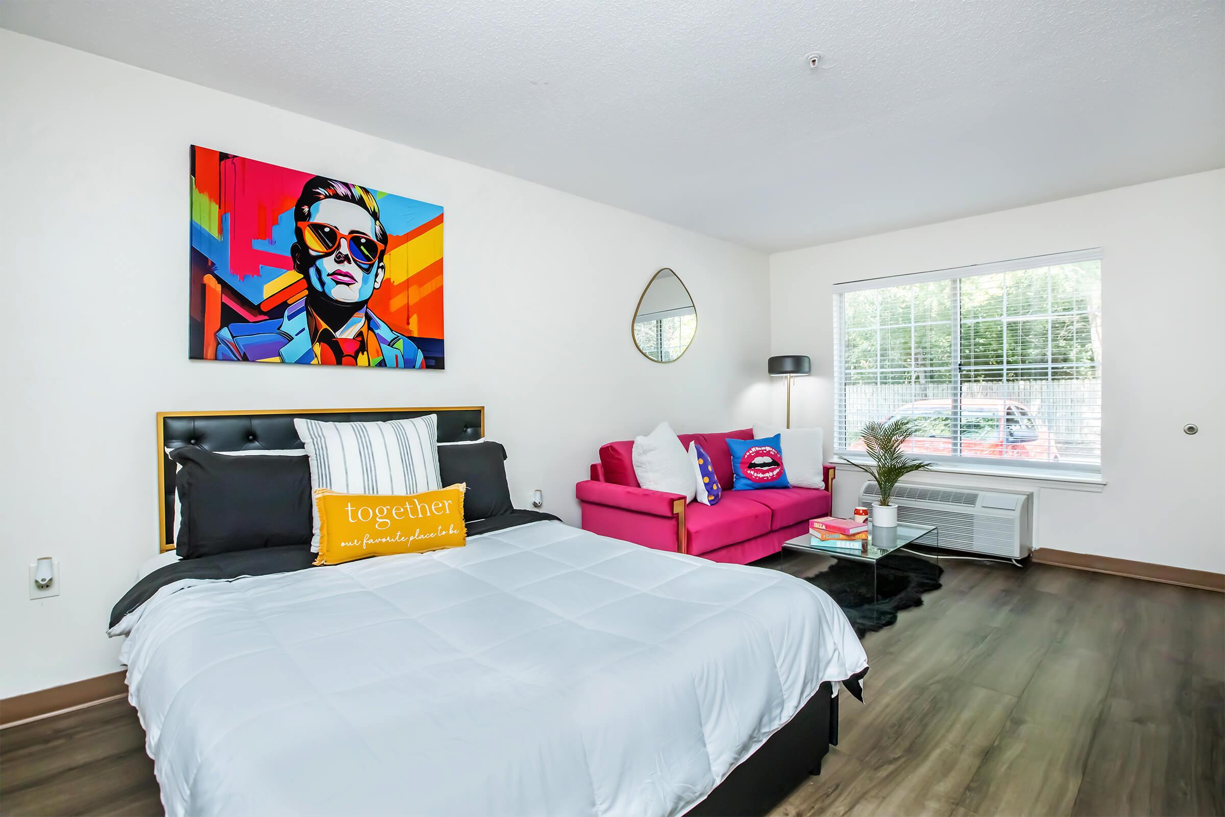A modern bedroom featuring a black bed with a white comforter, a vibrant pop art painting of a man in sunglasses on the wall, a pink sofa with decorative cushions, a round mirror, and a window with natural light. Minimalist décor and wooden flooring create a stylish atmosphere.