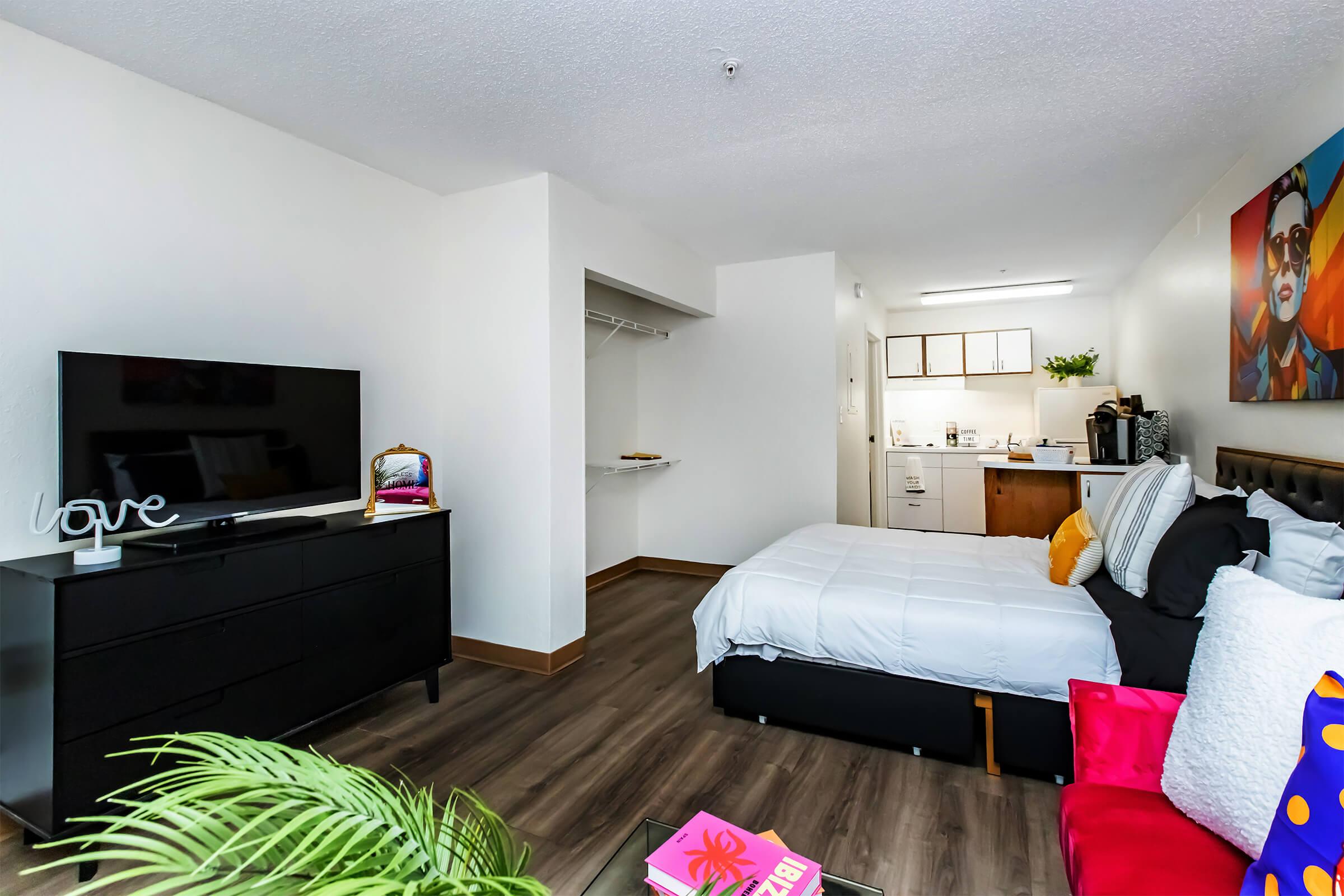 Cozy studio apartment featuring a bed, black dresser with a TV, and vibrant decor. A small kitchenette is visible, along with a seating area adorned with colorful cushions. Large window brings in natural light, enhancing the bright and modern atmosphere.