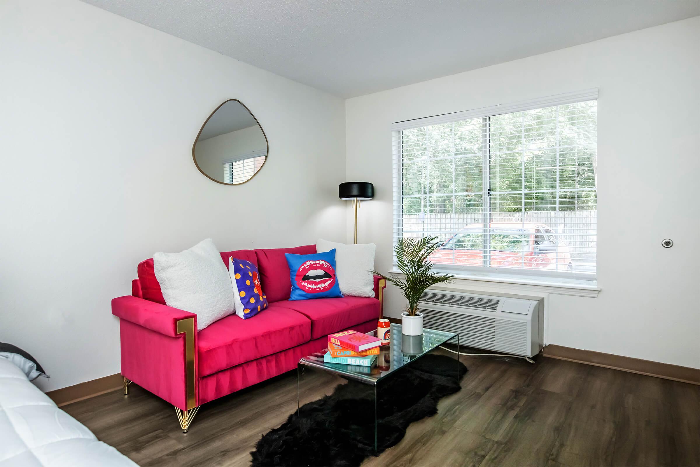 A vibrant living room featuring a bright pink sofa with colorful cushions, a modern glass coffee table, and a potted plant. Natural light streams in through a large window with blinds, and a stylish mirror hangs on the wall. An air conditioning unit is visible beneath the window, completing the cozy atmosphere.