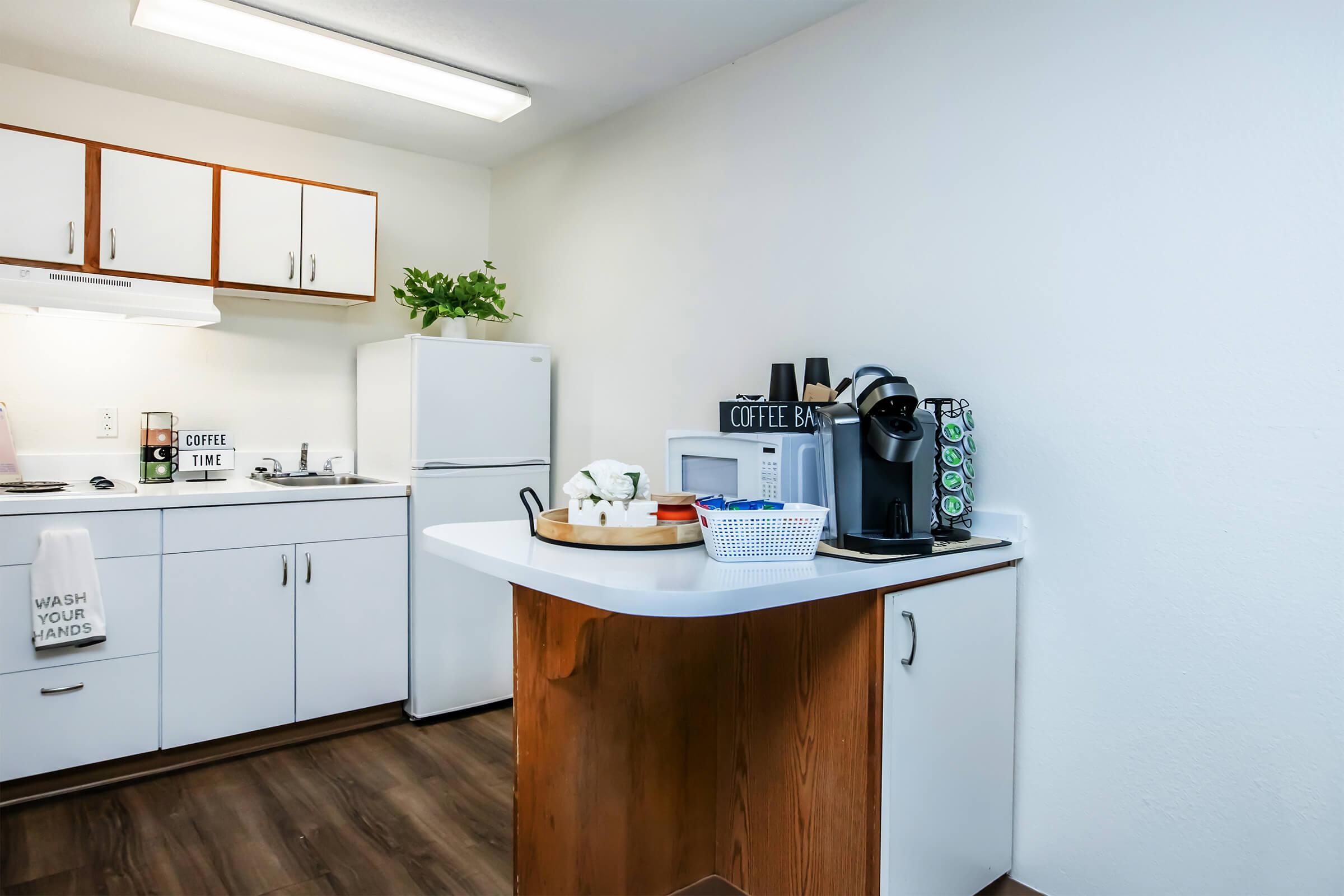 Bright and modern kitchen with white cabinets and appliances, including a refrigerator and microwave. A small island counter with a coffee maker, decorative items, and a basket is featured. A plant adds a touch of greenery to the space. A towel with the phrase "WASH YOUR HANDS" is hanging by the sink.