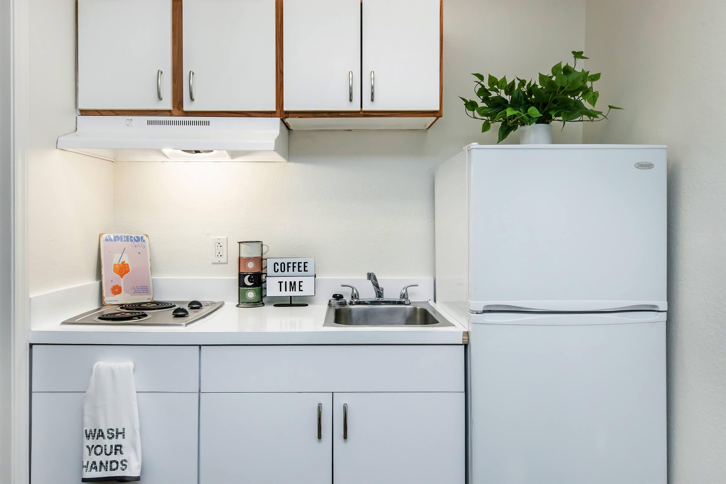 A small, modern kitchen featuring white cabinets and appliances. There's a sink, a stove with a hood, and a refrigerator. A decorative plant sits on top of the fridge. A sign reads "COFFEE TIME," and a towel is hanging by the sink, stating "WASH YOUR HANDS." Natural light brightens the space.