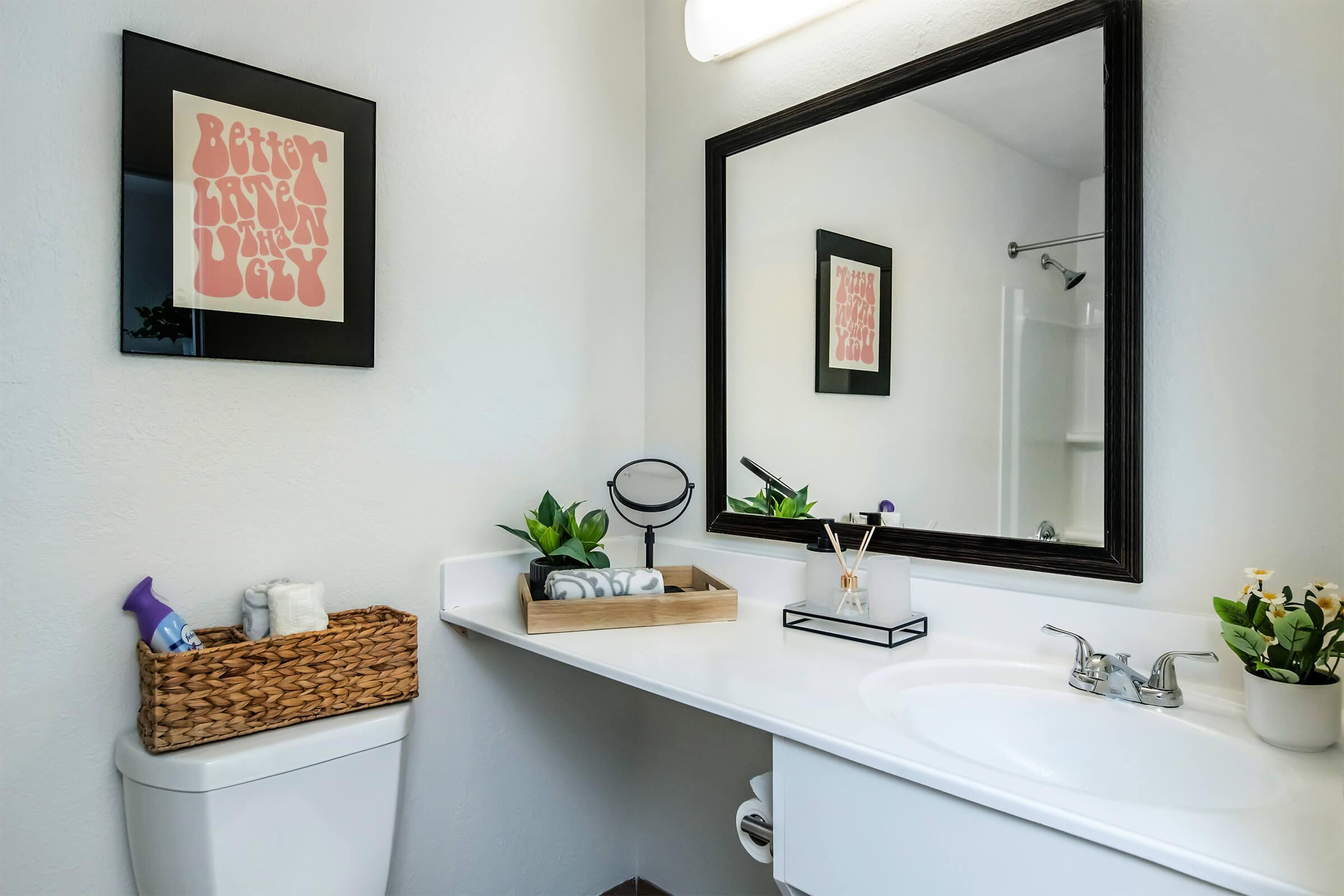 A modern bathroom featuring a large mirror above a white countertop. There is a decorative black-framed art piece on the wall, alongside potted plants. A woven basket rests on the counter, containing cleaning supplies and toiletries. The overall decor is minimal and fresh, with light-colored walls.